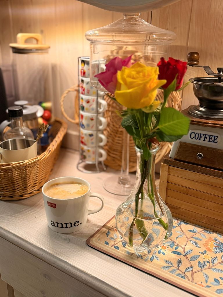 A warm, inviting home beverage bar featuring a ceramic mug of frothy latte text reading anne, a clear glass vase with fresh yellow and red roses, a wooden vintage coffee grinder, and woven wicker storage baskets on a light wood counter.