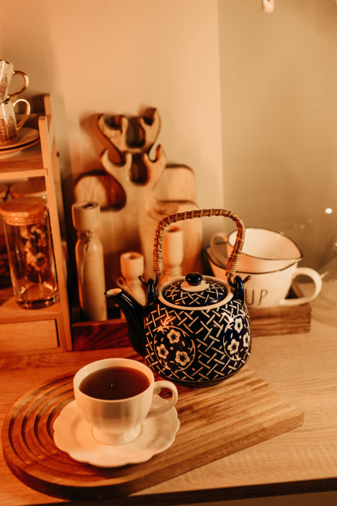 A blue and white lattice-patterned ceramic teapot with a woven bamboo handle sitting next to a matching cup of black tea on a grooved wooden serving board in a warm kitchen setting.