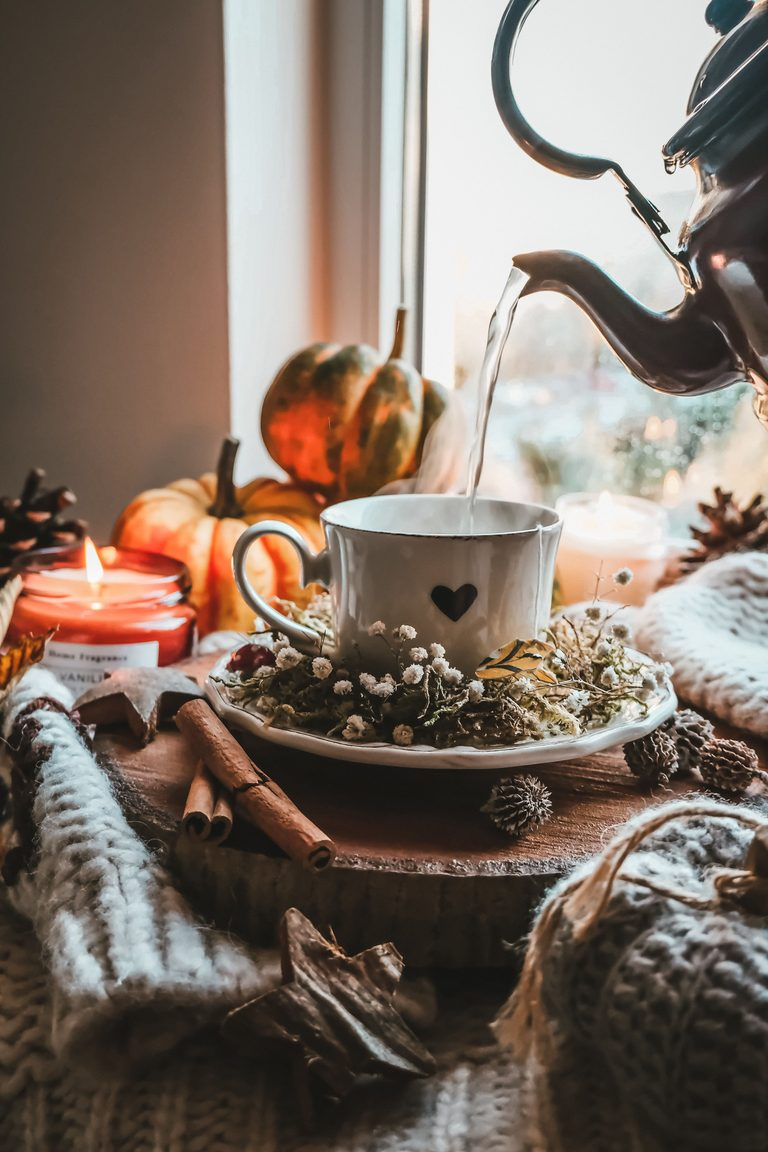 An aesthetic autumn scene for a tea lovers gift guide featuring hot water pouring from a vintage copper kettle into a white ceramic heart teacup on a rustic wood slice surrounded by pumpkins and pinecones.