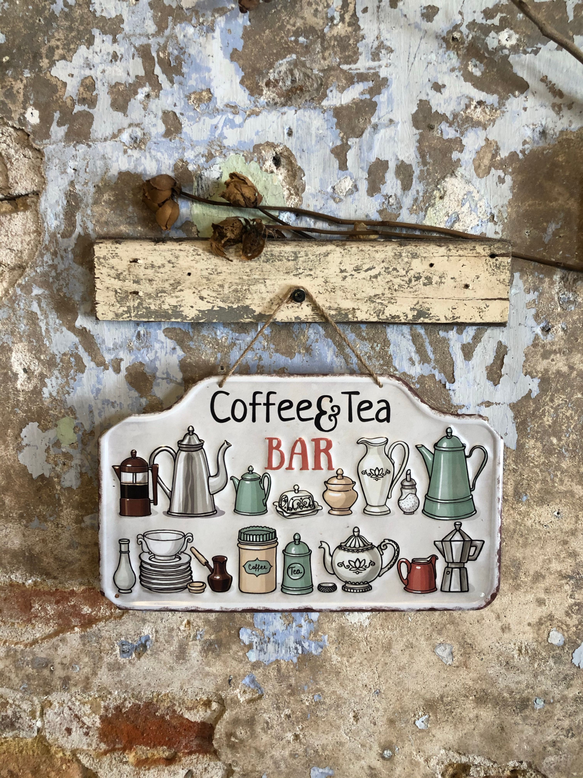 A rustic tin sign reading Coffee & Tea Bar with illustrations of vintage teapots, french presses, and espresso makers hanging on a weathered, peeling plaster wall beneath a piece of distressed wood and dried flowers.